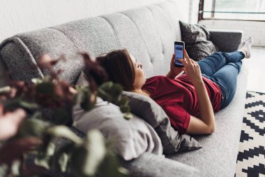 Evde kanepede yatan ise akıllı telefon kullanan genç kadın