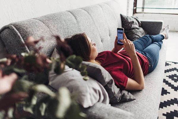 Evde kanepede yatan ise akıllı telefon kullanan genç kadın — Stok fotoğraf
