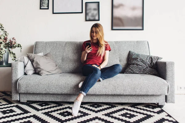 Mladá žena doma sedí na gauči, pomocí smartphonu — Stock fotografie