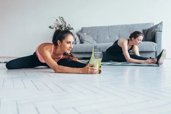 Deportivas calentando los músculos antes del entrenamiento haciendo ejercicio de estiramiento sentadas en el suelo en casa —  Fotos de Stock