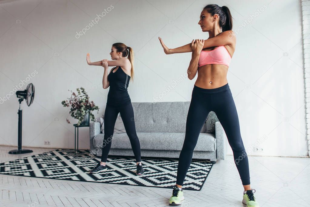 Two fit young woman stretching their arms exercising at home. Caucasian female fitness models working out in the morning.