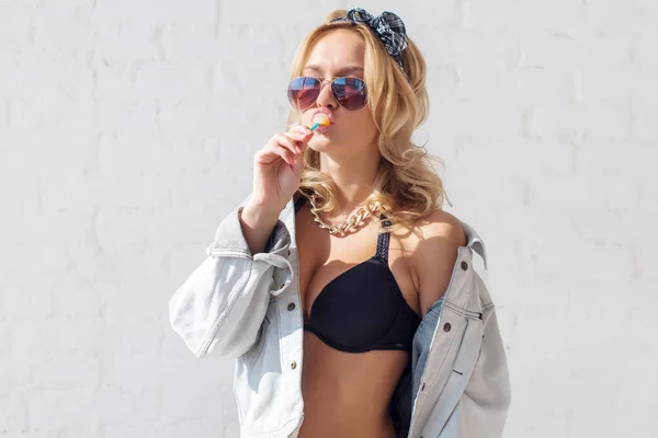 Junge sexy Frau mit Lutscher im Mund trägt Sonnenbrille BH und Jeansjacke im Freien, Lifestyle. — Stockfoto