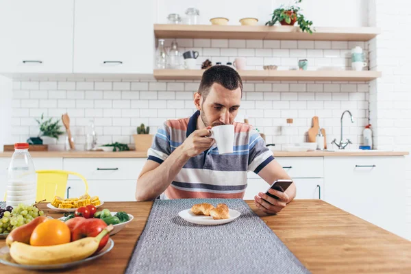 Jeune homme assis à table dans la cuisine avec une tasse de café à l'aide d'un téléphone mobile . — Photo