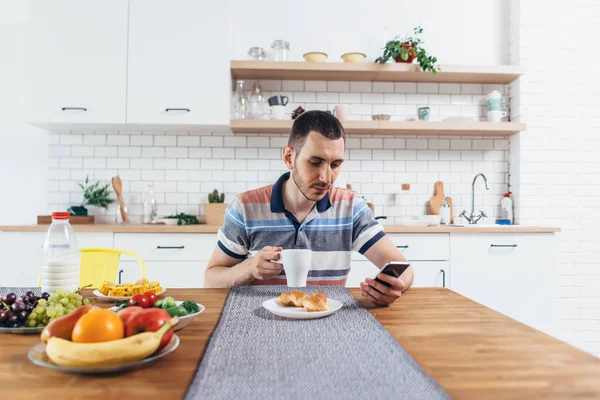 Młody człowiek za pomocą jego smartphone jedząc śniadanie w domu. — Zdjęcie stockowe