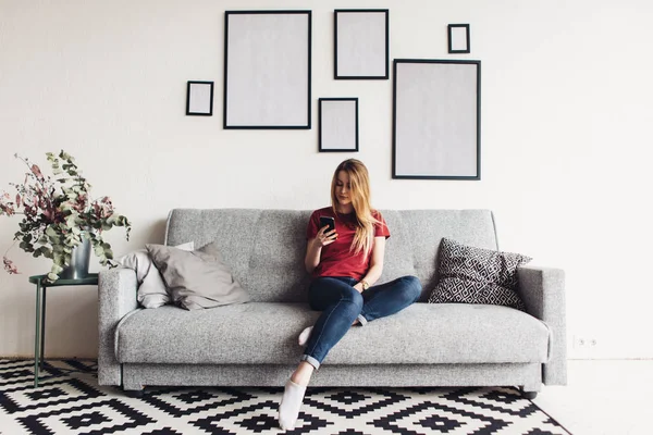 Junge glückliche Frau sitzt auf Sofa und benutzt Mobiltelefon zu Hause — Stockfoto