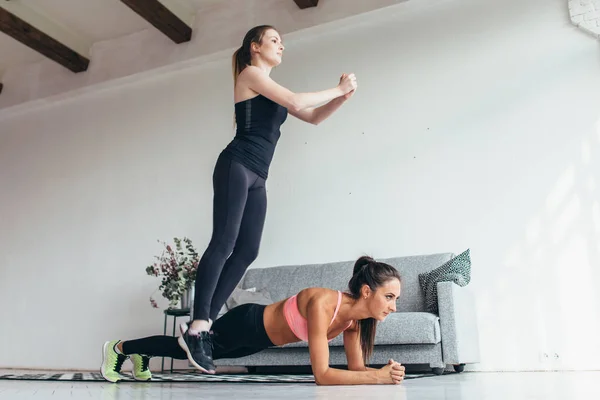 Mulheres aptas a treinar em casa. Menina pulando sobre seu amigo enquanto mulher realizando posição prancha . — Fotografia de Stock