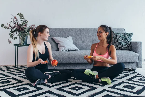 Sorridente donne in forma che indossano abbigliamento sportivo seduto sul pavimento mangiare frutta e parlare — Foto Stock
