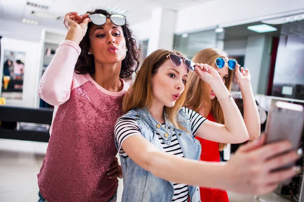 Tres novias jóvenes y elegantes que levantan gafas de sol de moda mientras se toman selfie con teléfono inteligente en el centro comercial —  Fotos de Stock