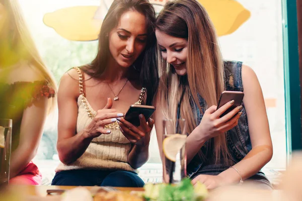 Две улыбающиеся кавказские подружки смотрят фото и видео на смартфоне, сидя в кафе и обедая — стоковое фото