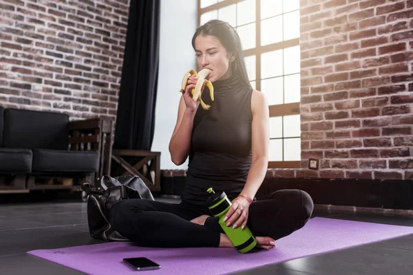 Porträtt av unga kaukasiska kvinna vila efter träning håller en sport flaska sitter på mattan med benen korsade i gymmet med loft interiör — Stockfoto