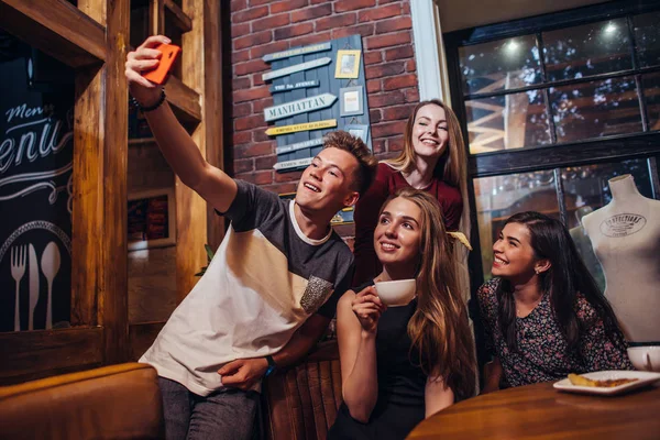 Group of smiling attractive teenagers wearing casual outfit taking selfie with mobile phone drinking tea in a cafe with loft interior — Stock Photo, Image