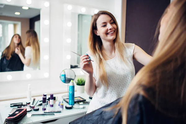 Joven artista de maquillaje alegre trabajando con un cliente sosteniendo un cepillo delgado de pie en el estudio de maquillaje — Foto de Stock