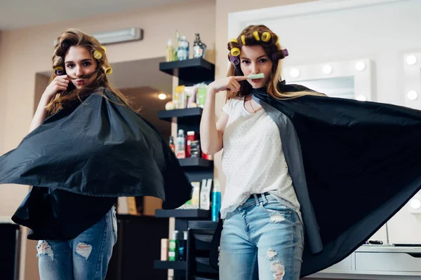 Two female friends goofing around in front of the camera making moustache from hair and curler standing in flying cape like heroes at beauty shop