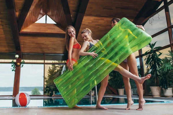 Attractive smiling slim female friends wearing swimsuits holding inflatable lounge posing in spa and wellness center — Stock Photo, Image
