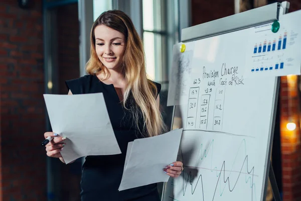 Female office worker working on her presentation standing near white board reading the report printed on paper — Stock Photo, Image