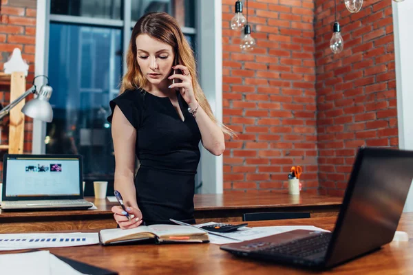 Mujer bastante joven hablando por teléfono utilizando una calculadora que trabaja en la oficina de pie en el escritorio — Foto de Stock