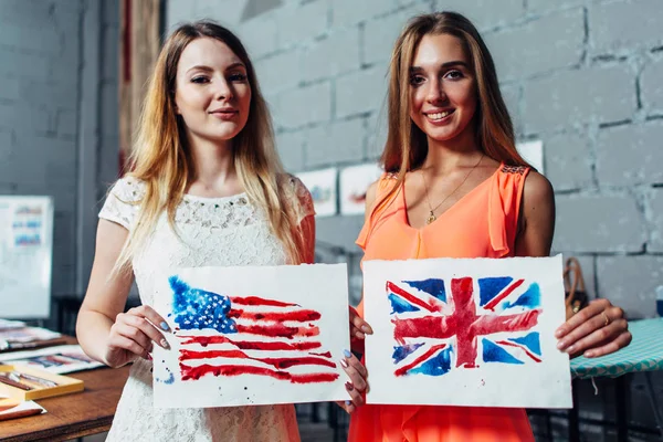 Close-up image of two young women holding a drawing of British and American flags hand-drawn with aquarelle technique on plain paper — Stock Photo, Image