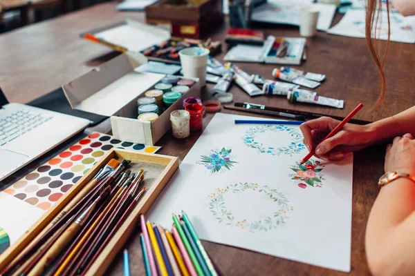 Zbliżenie: kobiece projektant rysunek kompozycje kwiatowe z kredki siedzi w miejscu pracy, otoczony z farby, gwasz, pędzle oraz inne sztuki dostarcza — Zdjęcie stockowe