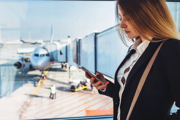 Succesvolle jonge vrouwelijke freelancer permanent in de buurt van venster op de luchthaven een SMS van de telefoon te houden — Stockfoto