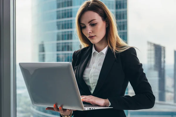 Unga kvinnliga jurist som arbetar i hennes lyxiga kontor håller en laptop står mot panoramafönster med utsikt över affärsdistriktet — Stockfoto