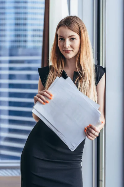 Portrait of pretty smiling young woman holding papers standing at window with cityscape view — Stock Photo, Image