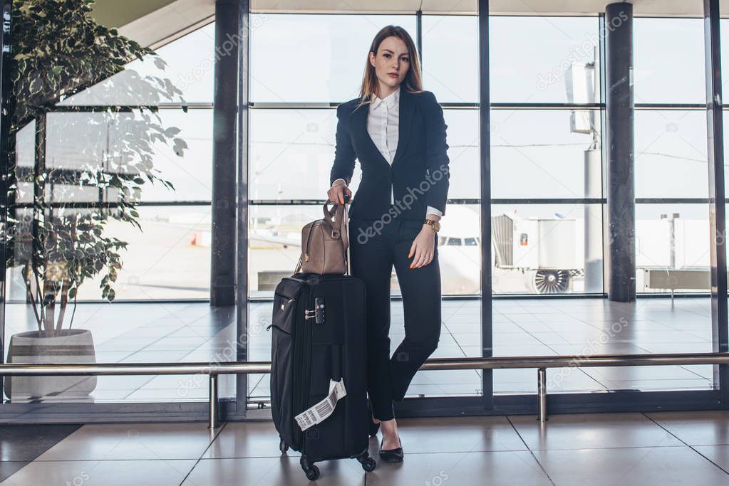 Full-length portrait of confident young business traveler wearing formal suit standing with heavy roll-aboard suitcase in airport terminal