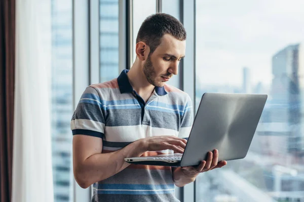 Бизнесмен, держащий в руках ноутбук, стоящий в офисе у окна с видом на город — стоковое фото