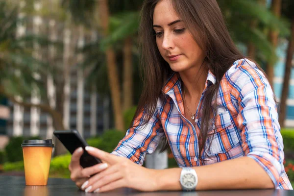 Mujer joven sentada al aire libre leyendo y escribiendo mensajes en su teléfono inteligente . — Foto de Stock