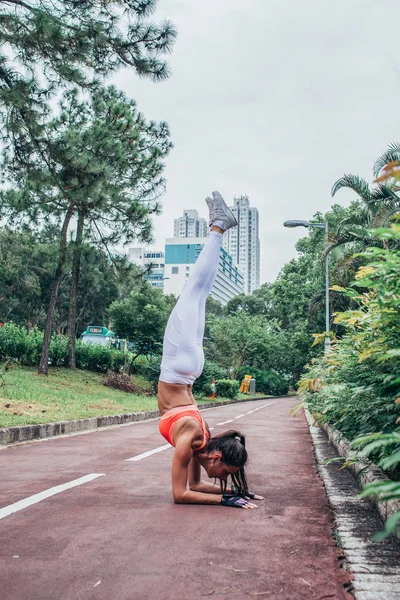 Sportswoman doing handstand yoga exercise standing on her forearms with straight legs path in the park view of city buildings