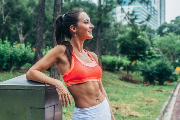 Schlanke Fitness-Brünette mit Sixpack-Bauch in rosa Sport-BH steht nach dem Training entspannt im Stadtpark und schaut von der Kamera weg — Stockfoto