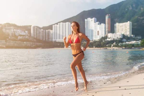 适合女运动员穿着比基尼在海滩上运行阳光照耀在相机与背景在山酒店度假村 — 图库照片