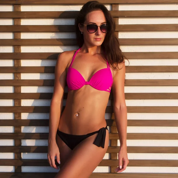 Närbild foto av garvade brunett modell med sexig kropp står i bikini och solglasögon mot vägg trä galler på het sommar dag solen skiner ljust — Stockfoto