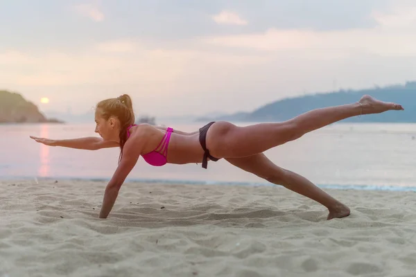 Mayo kumsalda bacaklarını deniz günbatımı sırasında germe egzersiz genç kadın. Fitness kız sahil üzerinde yapıyor — Stok fotoğraf