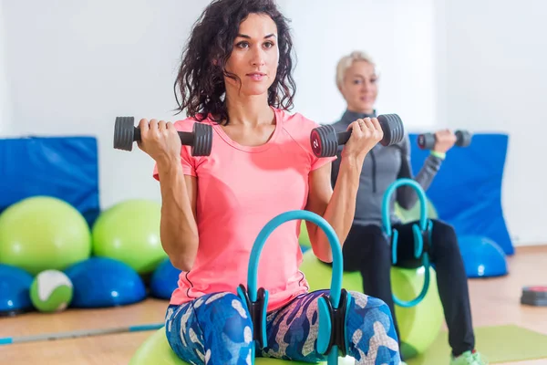 Schlanke, sportliche Frauen, die auf Gymnastikbällen sitzen, Hanteln in der Hand halten und Pilates-Ring zwischen die Beine pressen während des Gruppenkurses im Fitnessstudio — Stockfoto