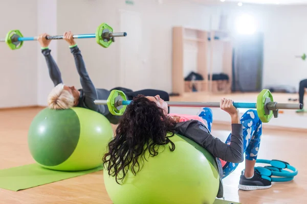 Задний вид двух женщин, поднимающих штангу, лежащих на мяче стабильности во время тренировки в спортзале — стоковое фото
