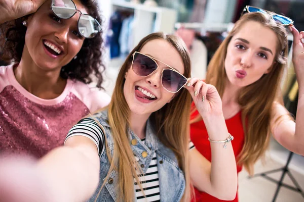 Las novias sonrientes que usan gafas de sol elegantes que se divierten tomando selfie con el teléfono móvil mientras que hacen compras en la tienda de ropa —  Fotos de Stock