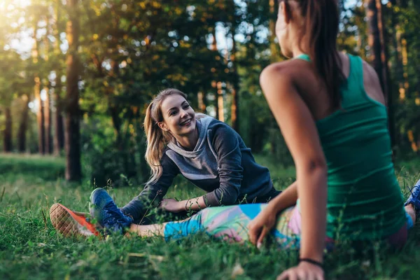 Two cheerful female athletes warming up before training doing stretching exercise for legs sitting on grass — Stock Photo, Image