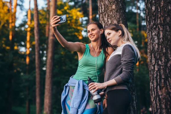 Zwei junge sportliche Freundinnen in Sportbekleidung lehnen an Baum und machen Selfie mit Smartphone im Wald — Stockfoto