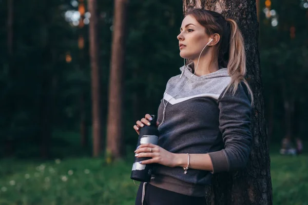 Portrait of pretty fit sportswoman wearing tracksuit leaning against tree listening to music in earphones holding sports water bottle standing in forest — Stock Photo, Image