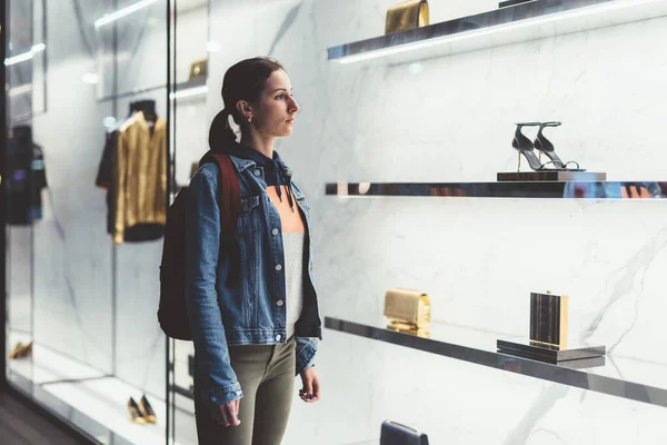 Ung kvinna står nära skyltfönster i butiken, tittar på handväskor och skor — Stockfoto