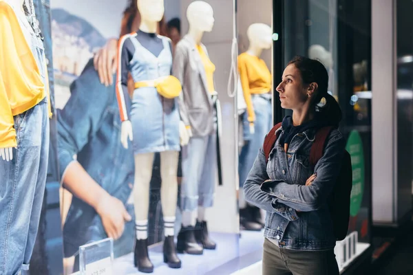 Fille se tient devant le magasin et regarde les vêtements féminins sur les mannequins. — Photo