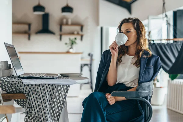 Kvinna som sitter vid köksbordet med bärbar dator och dricker ur en mugg — Stockfoto