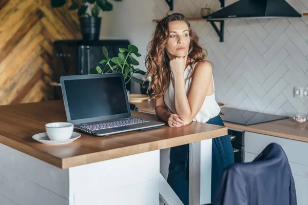 Pensive woman in the kitchen at the table with a laptop — Stock Photo, Image