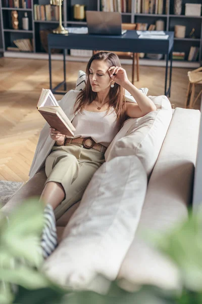 Jovem mulher em casa deitada no sofá e livro de leitura . — Fotografia de Stock