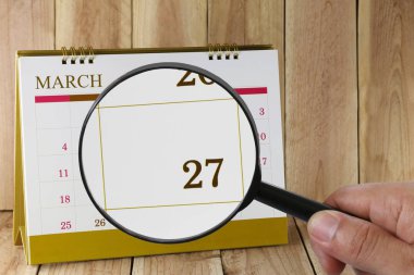 Magnifying glass in hand on calendar you can look Twenty seven day of month,Focus number Twenty seven in March,Concept in business and meetings. clipart