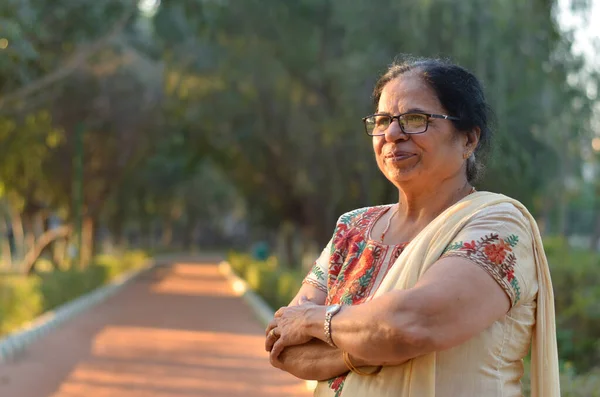 Smart and confident senior north Indian woman standing, posing for the camera with hands crossed / folded in a park wearing an off white salwar kameez punjabi suit in summers in New Delhi, India