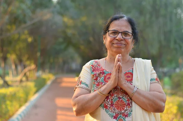 Smart senior north Indian woman standing, posing for the camera with hands folded in namaste as a sign of respect and welcome in a park wearing off white salwar kameez in summers in New Delhi, India