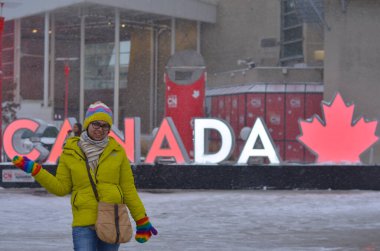 Young Indian woman posing in front of the lit up Canada 3d cut out sign installed in downtown Toronto, Ontario, Canada near Rogers center, CN tower and Ripley's aquarium on a snowy day clipart