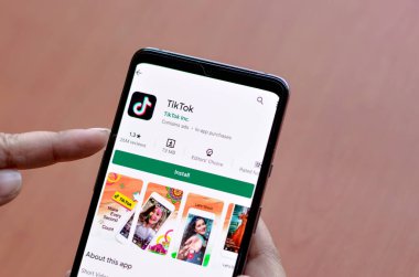 Yeni Delhi, Hindistan, 2020. TikTok mobil uygulamasını akıllı telefon ekranına yükleyen kadın, hızla büyüyen popüler sosyal ağ sitesinin logosunu gösteren ahşap arka planda, düşük reytingli
