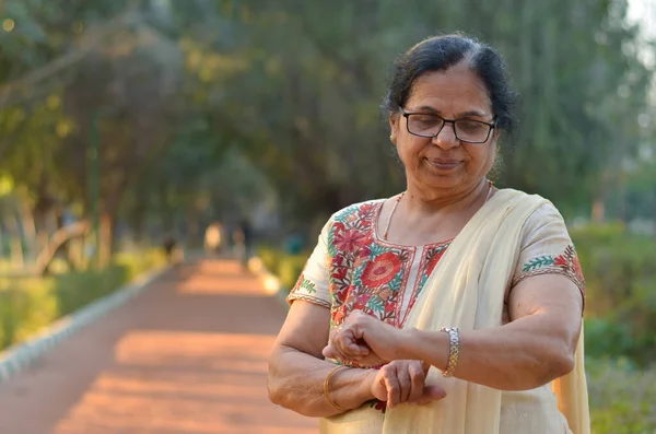 Senior Indian woman smiling and looking at her hands / smart watch to know the time in a park. She\'s wearing an off white salwar kamiz. Concept - Digital literacy in India for senior citizens
